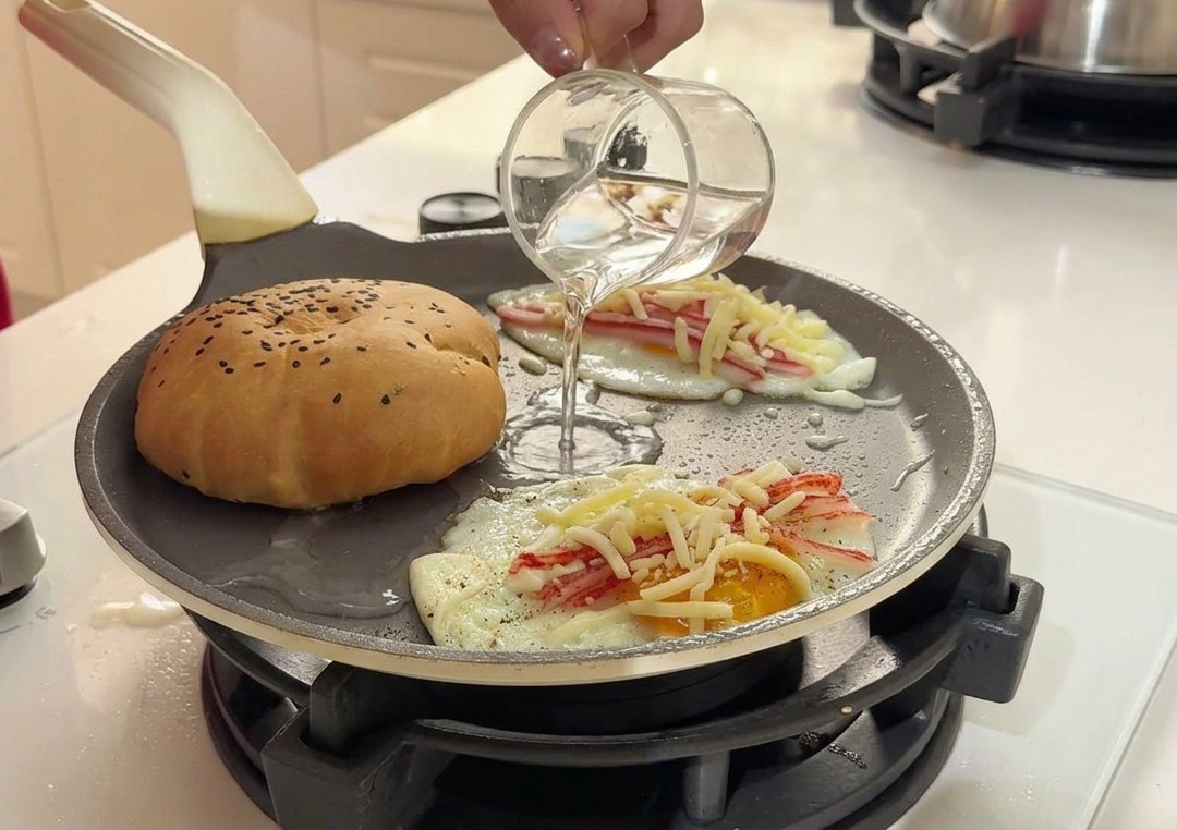 Vertiendo agua limpia desde un pequeño vaso de cristal a la sartén caliente entre el pan y los huevos.