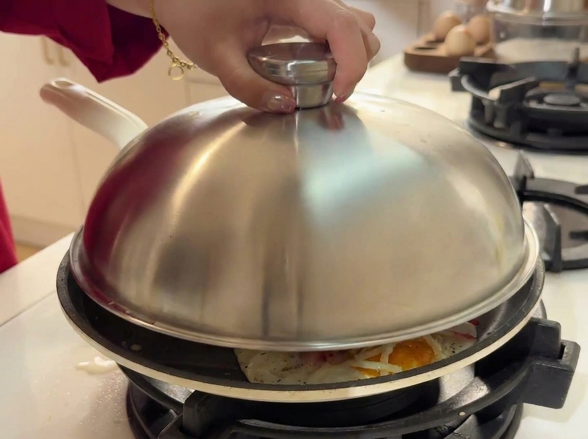 Una mano colocando una tapa de acero inoxidable sobre la sartén que contiene huevos, queso y palitos de cangrejo.