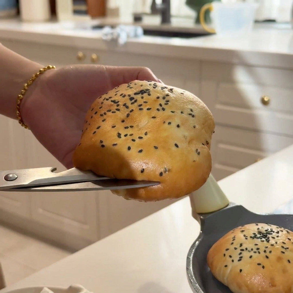Una mano cortando con cuidado un pan de sésamo caliente usando unas tijeras de cocina plateadas.