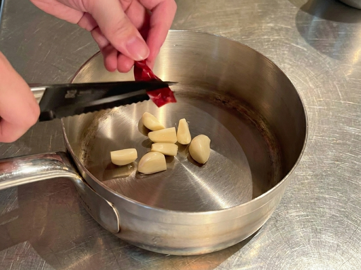Manos usando tijeras de cocina para cortar un chile rojo seco sobre la olla con ajo.