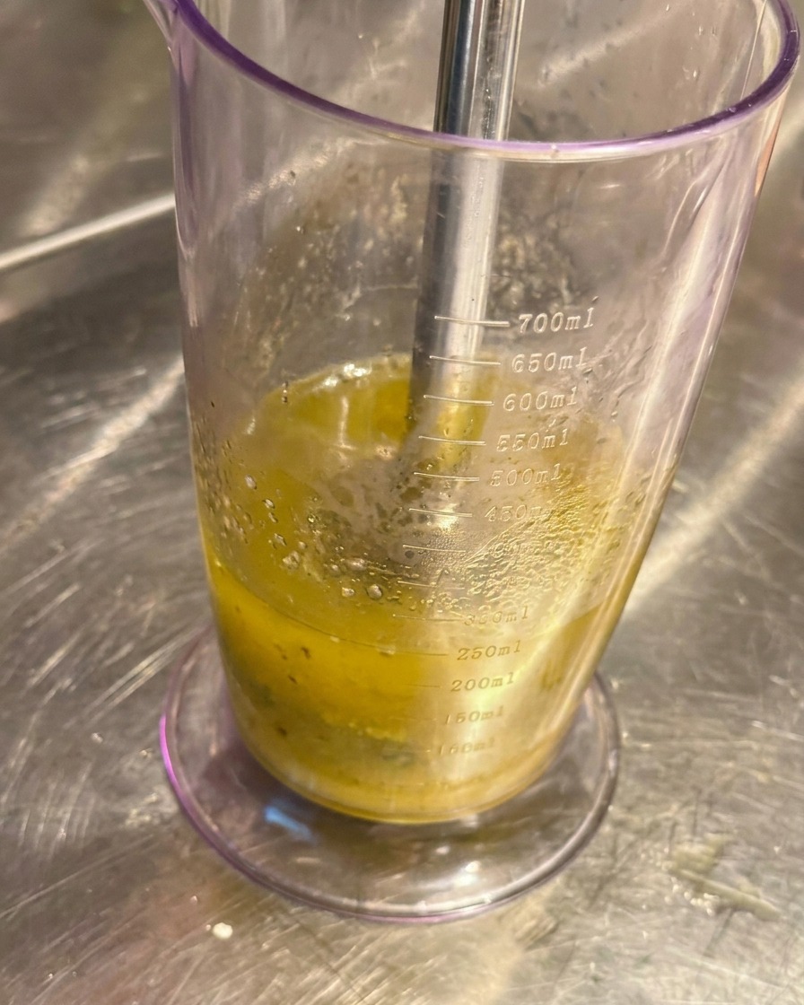 Batidora de mano en un vaso medidor triturando ajo confitado y aceite hasta formar una pasta pálida.