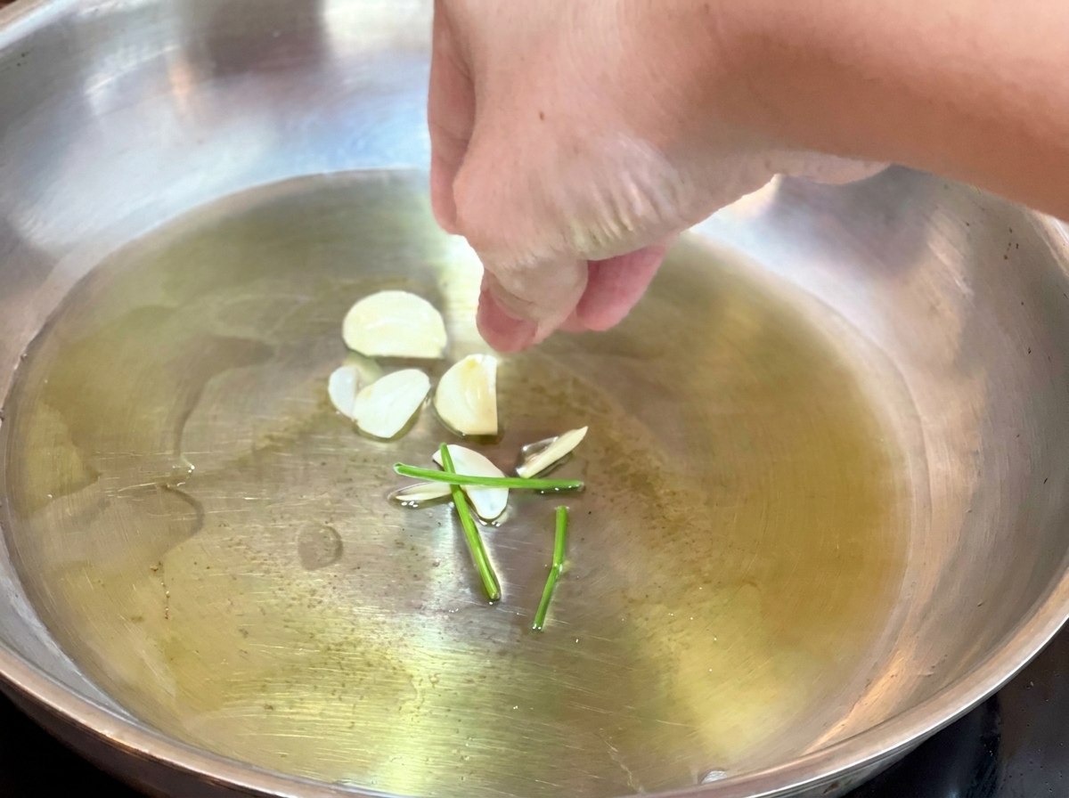 Mano echando láminas de ajo crudo y tallos de perejil en aceite caliente en una sartén.