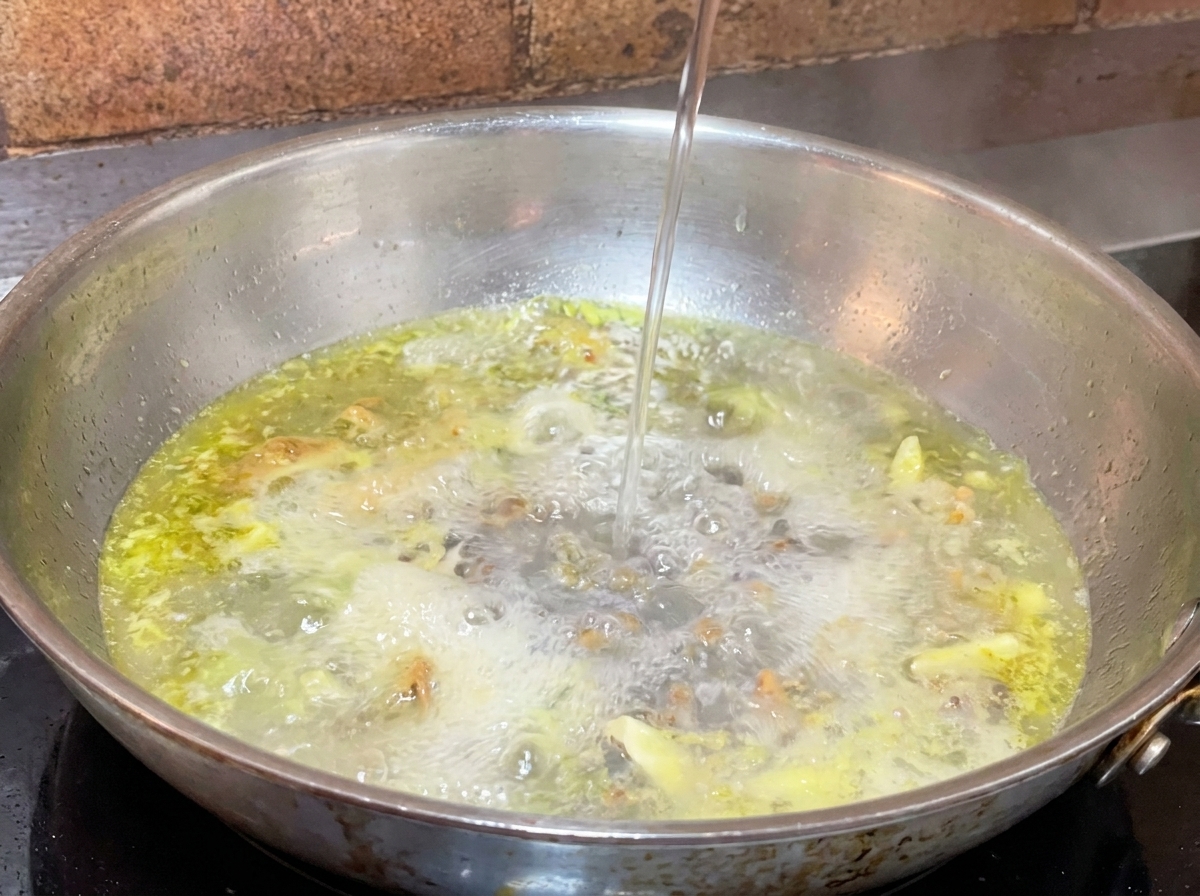 Chorro de agua de cocción de la pasta vertiéndose en una sartén con aceite y ajo burbujeante.