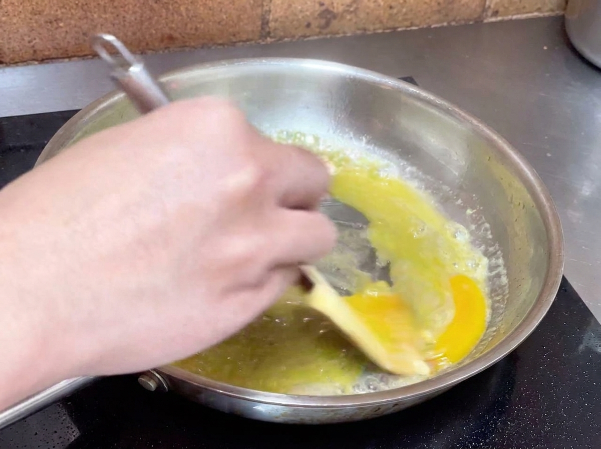 Mano removiendo enérgicamente una mezcla de aceite y agua en una sartén para crear una salsa cremosa.