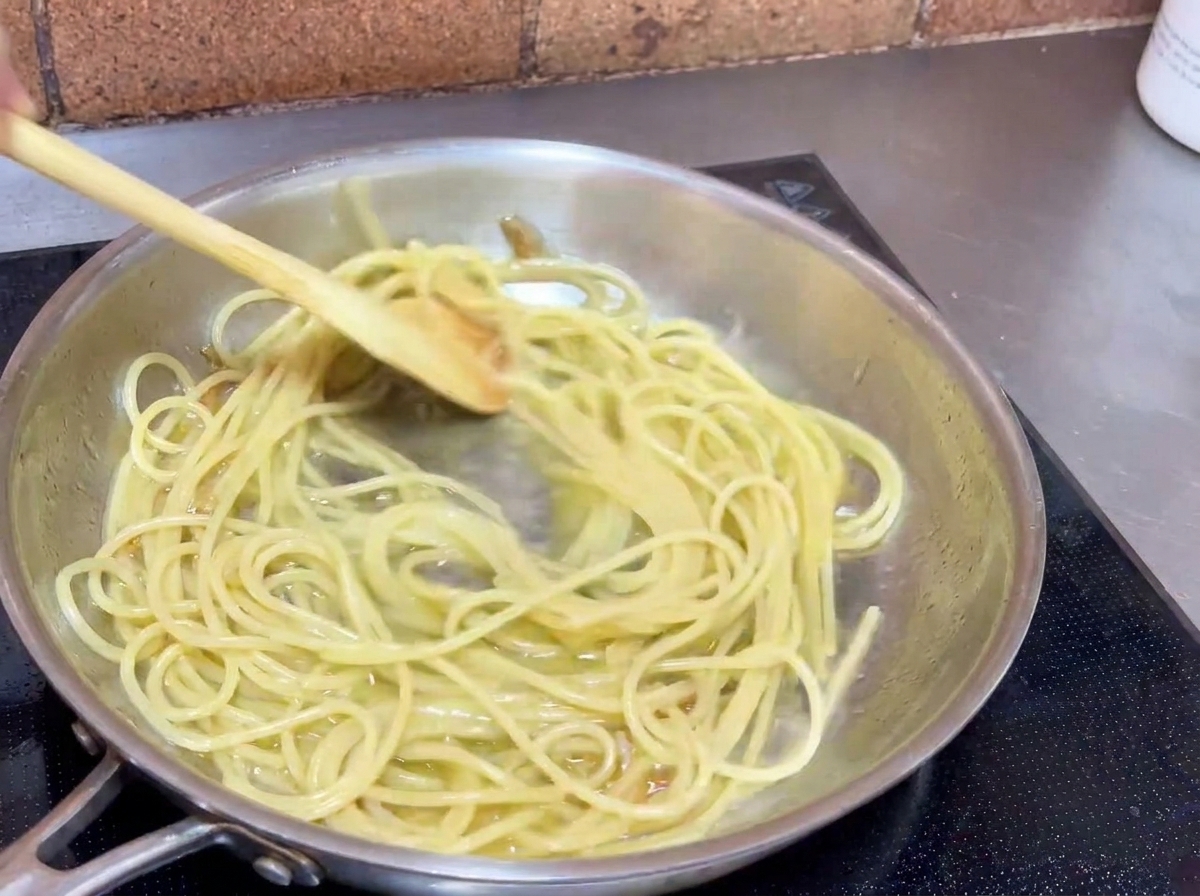 Espaguetis siendo removidos en una sartén con una cuchara de madera para cubrirlos con la salsa.