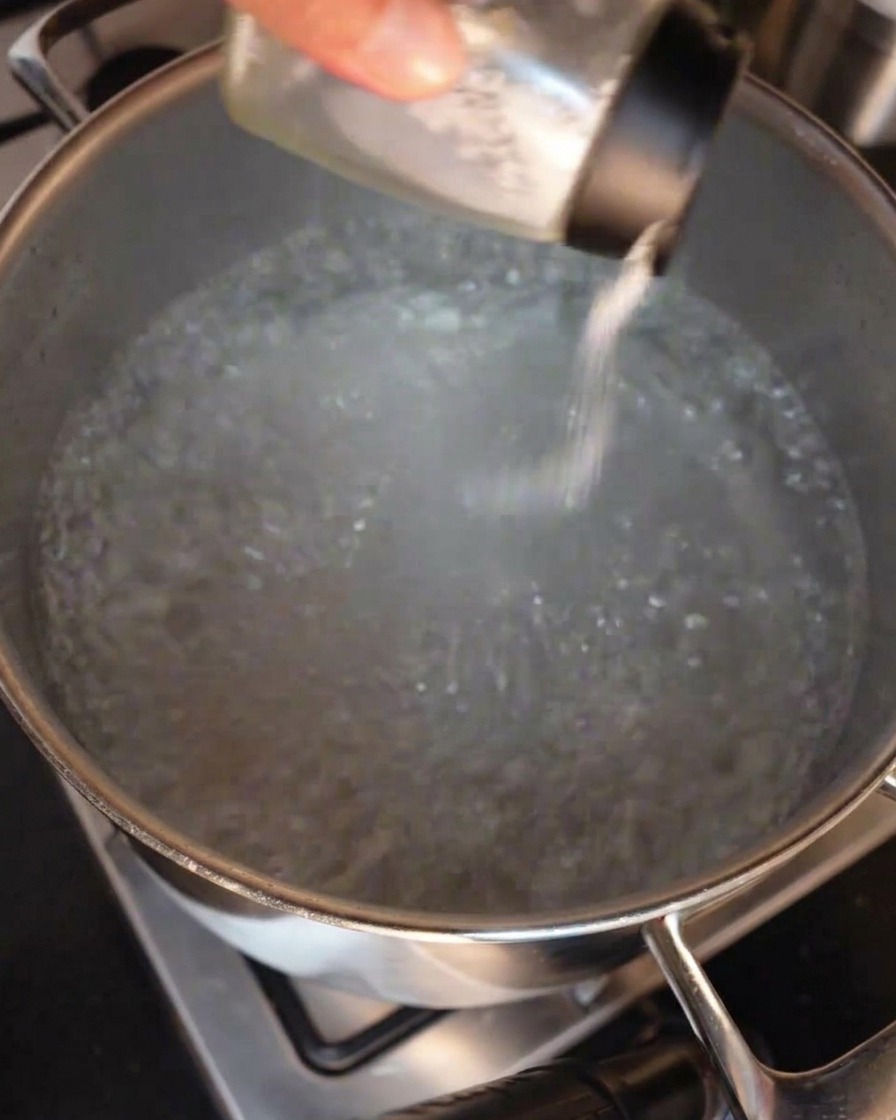 Una mano espolvoreando sal desde un salero de cristal en una olla grande de acero inoxidable con agua hirviendo a borbotones.