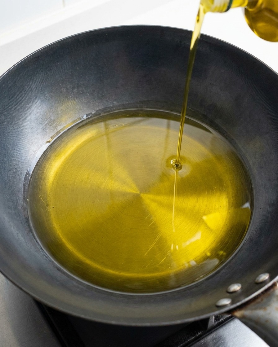 Un chorrito constante de aceite de oliva dorado vertiéndose en el centro de un wok o sartén oscura y seca.