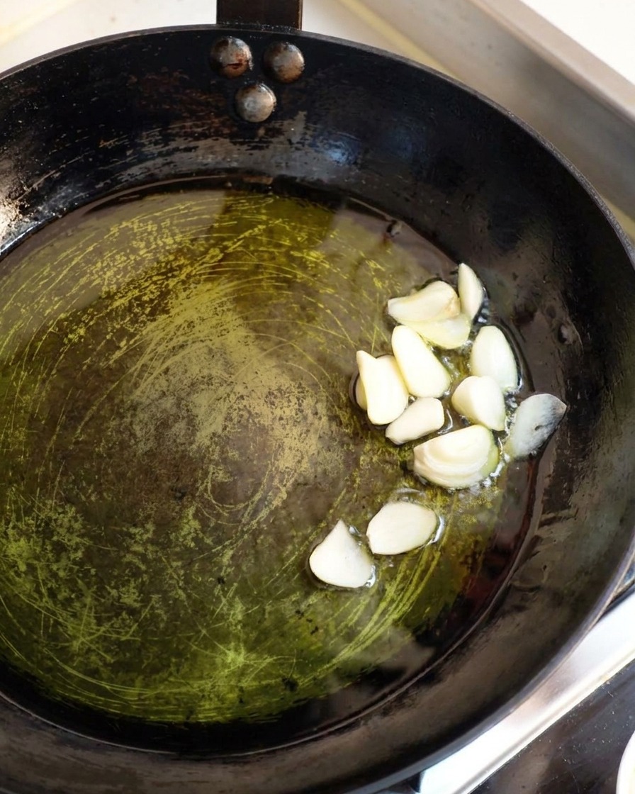 Gruesas láminas de ajo blanco crudo cocinándose a fuego lento en un charco poco profundo de aceite de oliva caliente dentro de una sartén oscura.