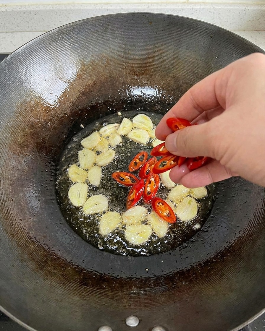 Una mano echando chiles rojos recién cortados en un wok con aceite caliente que ya está burbujeando con láminas de ajo.