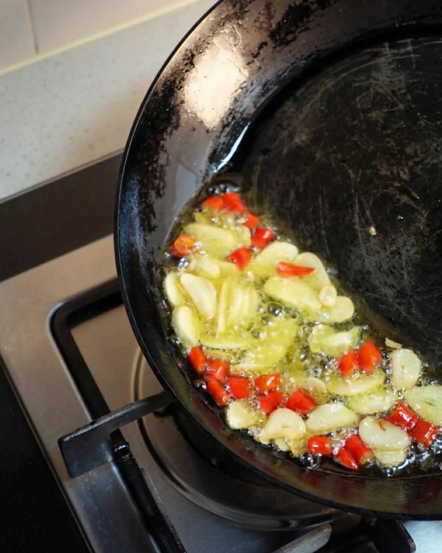 Un wok oscuro inclinado hacia un lado, acumulando aceite dorado caliente donde las rodajas de ajo y los chiles rojos picados burbujean y se fríen juntos enérgicamente.