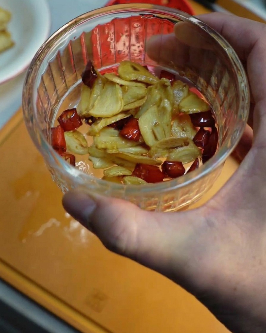 Una mano sosteniendo un pequeño bol de cristal lleno de crujientes láminas de ajo frito y trozos de chile rojo.