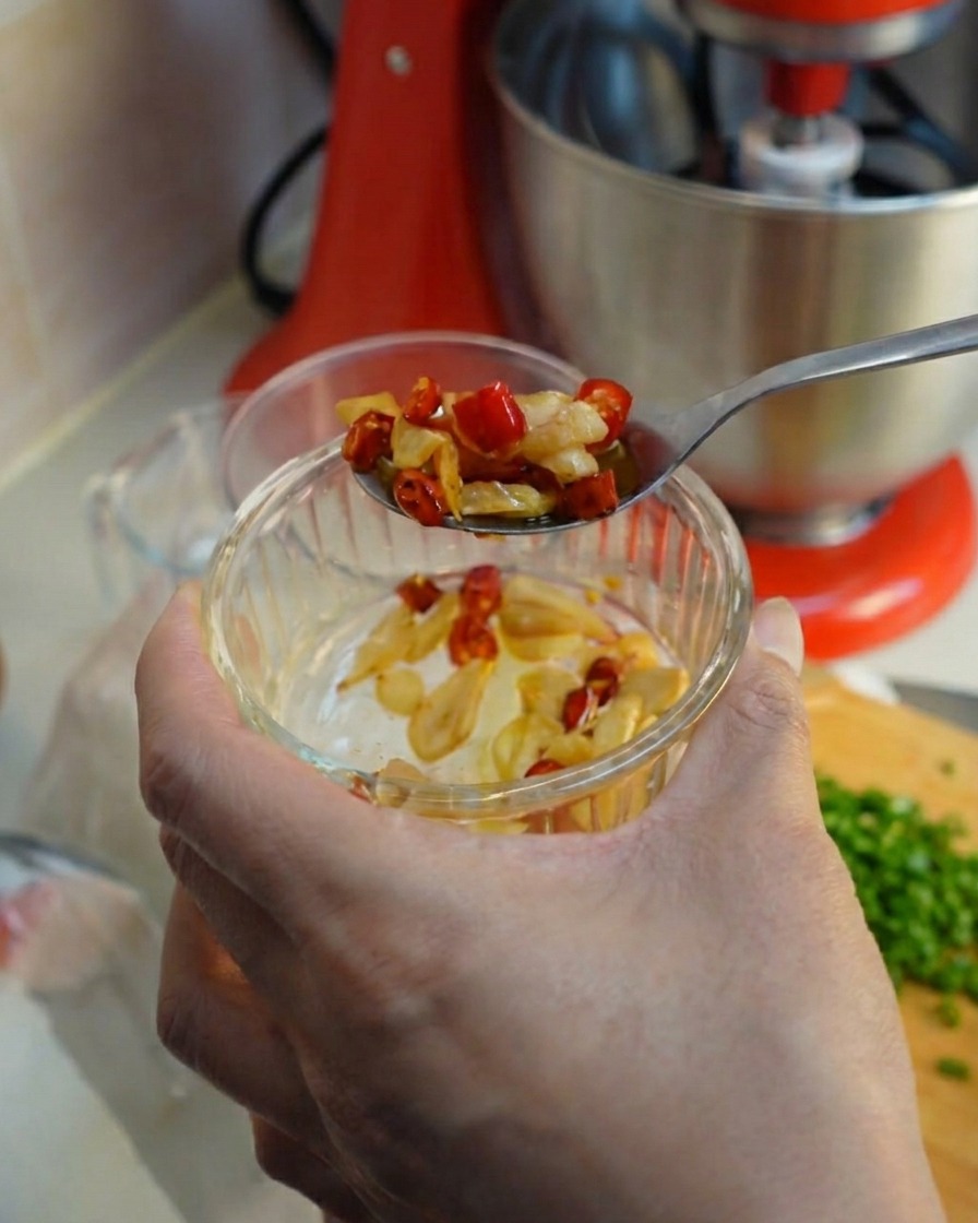 Una cuchara sacando ajo frito y rodajas de chile rojo de un pequeño bol de cristal.