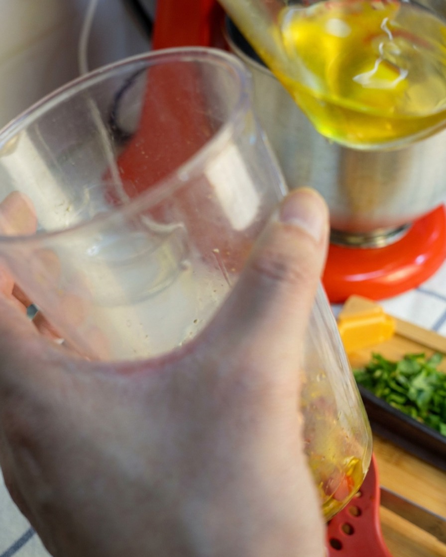Una batidora de mano levantándose de un vaso transparente, goteando con una salsa triturada suave y rojo dorada de ajo y chile.