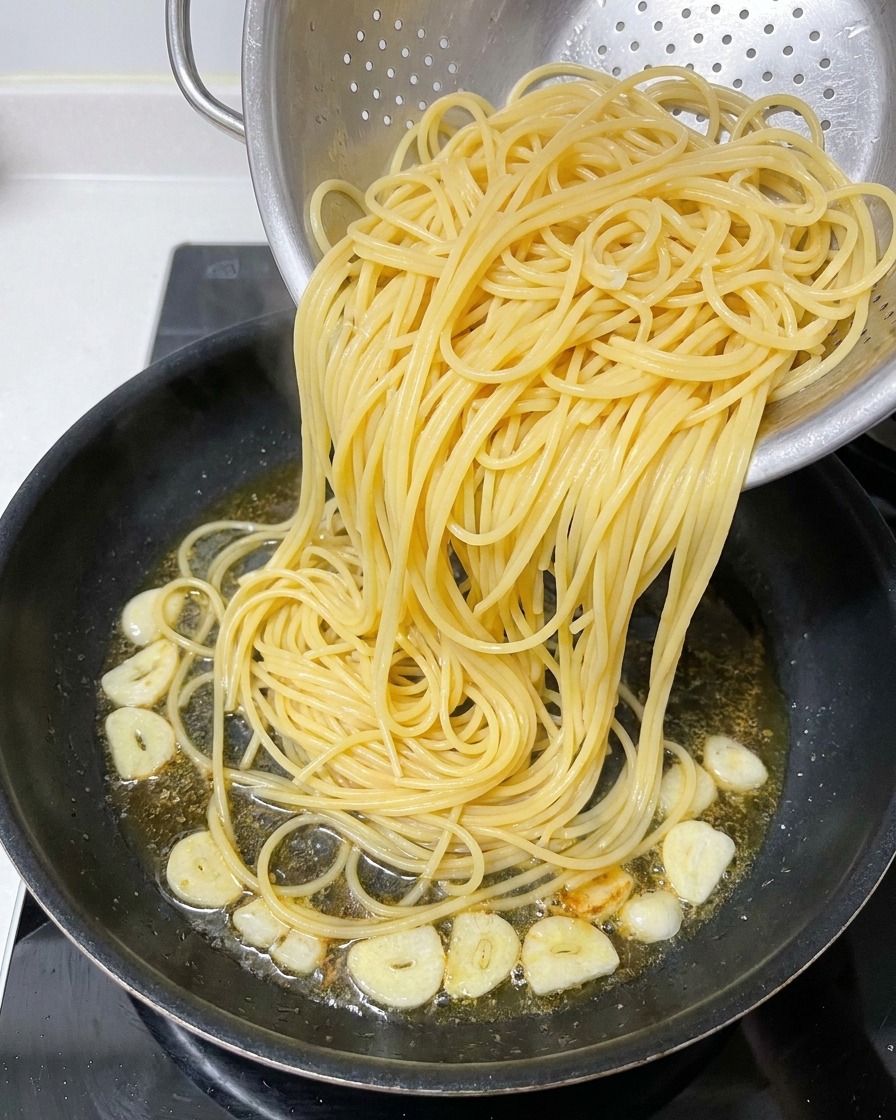 Espaguetis cocidos pasándose de un colador de metal a una sartén que contiene aceite infusionado y láminas de ajo frito.