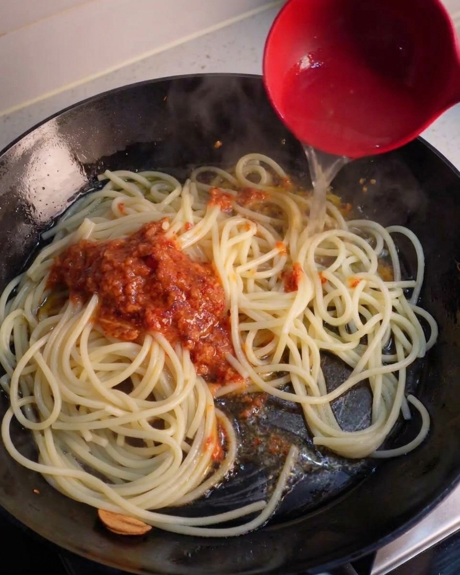Un cucharón rojo vertiendo agua de pasta transparente en una sartén sobre espaguetis y una cucharada de salsa triturada de ajo y chile rojo.
