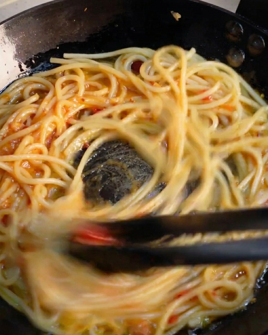 Pinzas negras salteando rápidamente espaguetis en una sartén para emulsionar la vibrante salsa de ajo y chile.