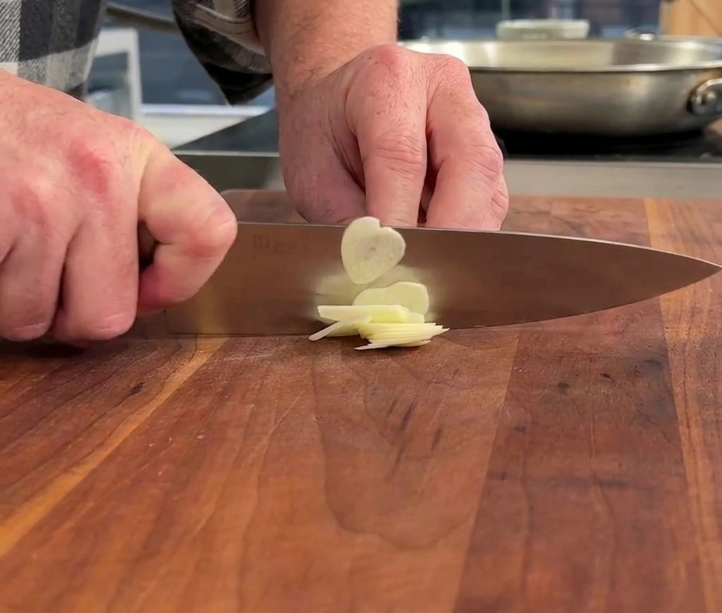 Manos usando un cuchillo de chef para cortar un diente de ajo en rodajas muy finas sobre una tabla de cortar de madera.
