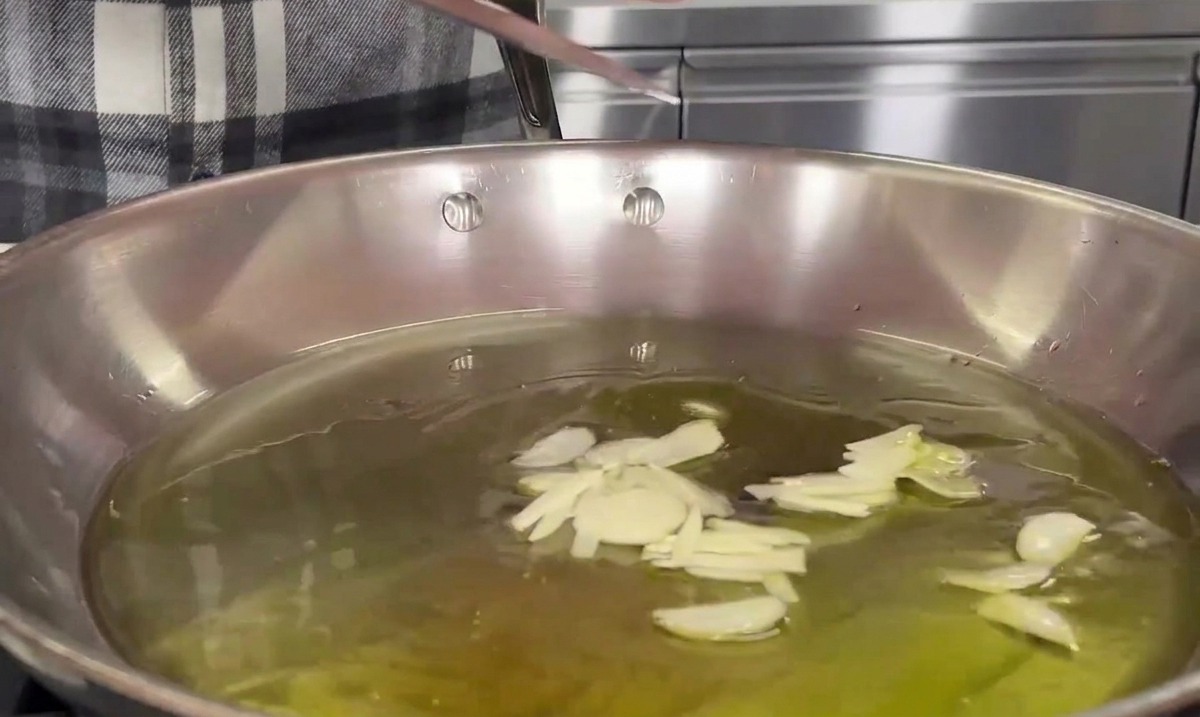 Usando la hoja de un cuchillo para deslizar ajo en rodajas finas en una sartén de acero inoxidable llena de aceite de oliva tibio.