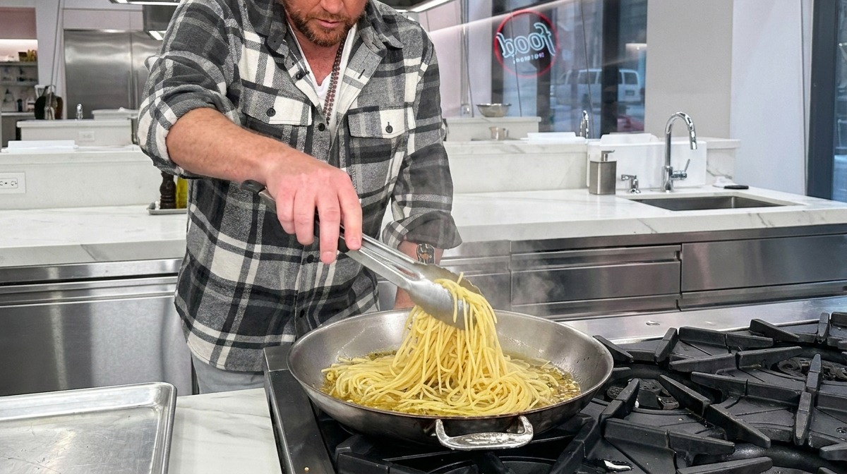 Espaguetis parcialmente cocidos cayendo en una sartén caliente con ajo, aceite y agua de pasta usando pinzas de metal.
