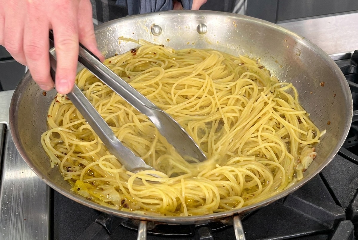Usando pinzas de metal para saltear espaguetis cocidos en una sartén de acero inoxidable con aceite de oliva, ajo y hojuelas de pimiento rojo.