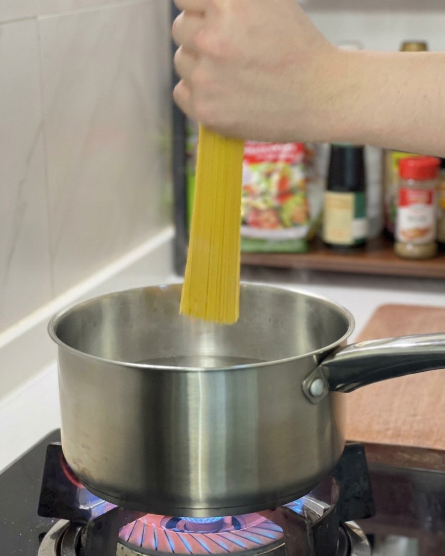 Mano sujetando un manojo de espaguetis secos introduciéndolos en una olla de acero inoxidable con agua hirviendo.