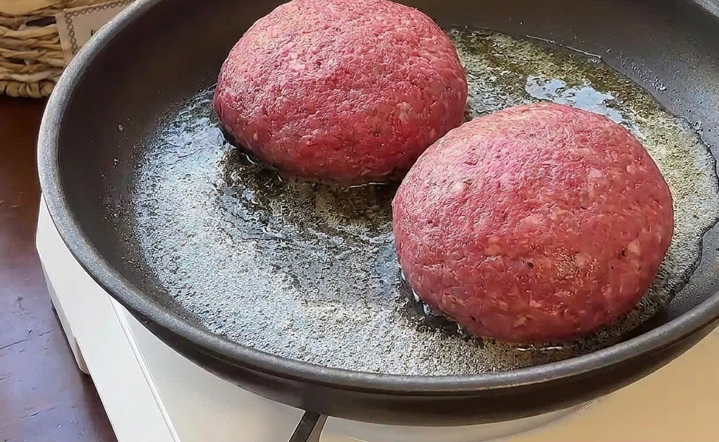 Dos hamburguesas gruesas de ternera cruda cocinándose en mantequilla derretida en una sartén portátil.