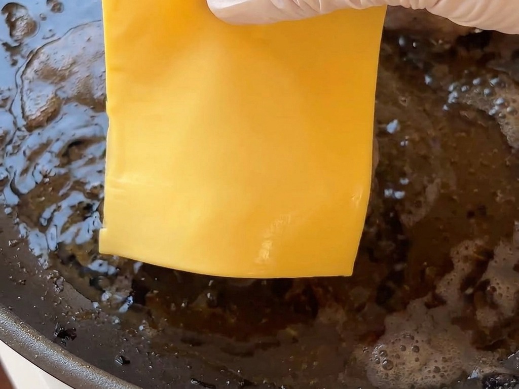 Una mano colocando una loncha cuadrada de queso sobre una hamburguesa caliente en la sartén.