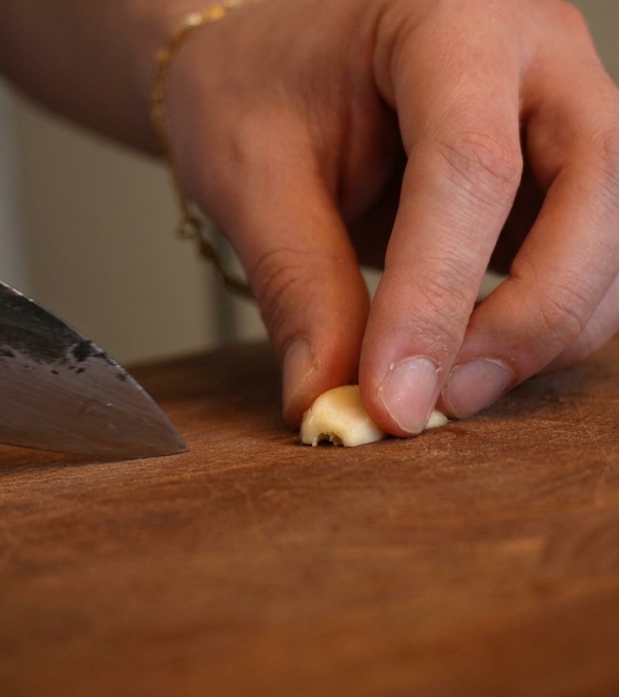 Primer plano de dedos sosteniendo un trozo de ajo mientras un cuchillo de chef lo pica finamente.