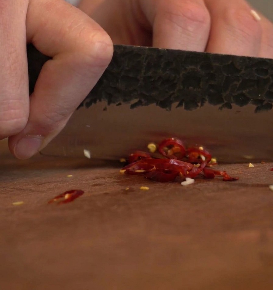 La hoja de un cuchillo oscuro picando chiles rojos secos sobre una tabla de madera.
