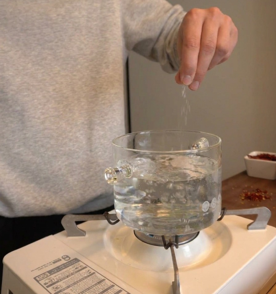 Mano espolvoreando sal en una olla de vidrio con agua hirviendo sobre una estufa portátil.
