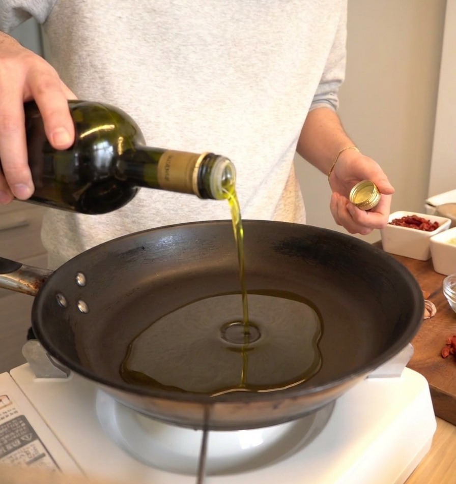 Aceite de oliva vertiéndose desde una botella verde oscuro en una sartén negra.