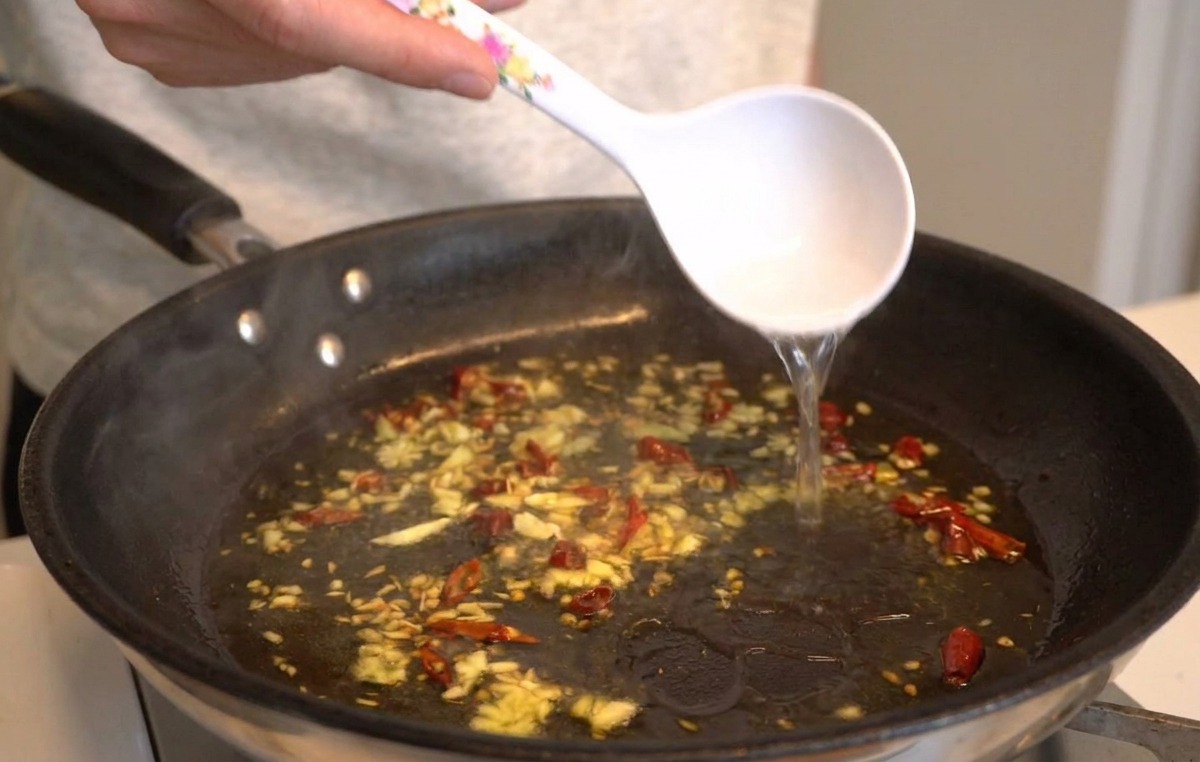 Un cazo blanco vertiendo agua caliente con almidón en una sartén con ajo, aceite y chiles.