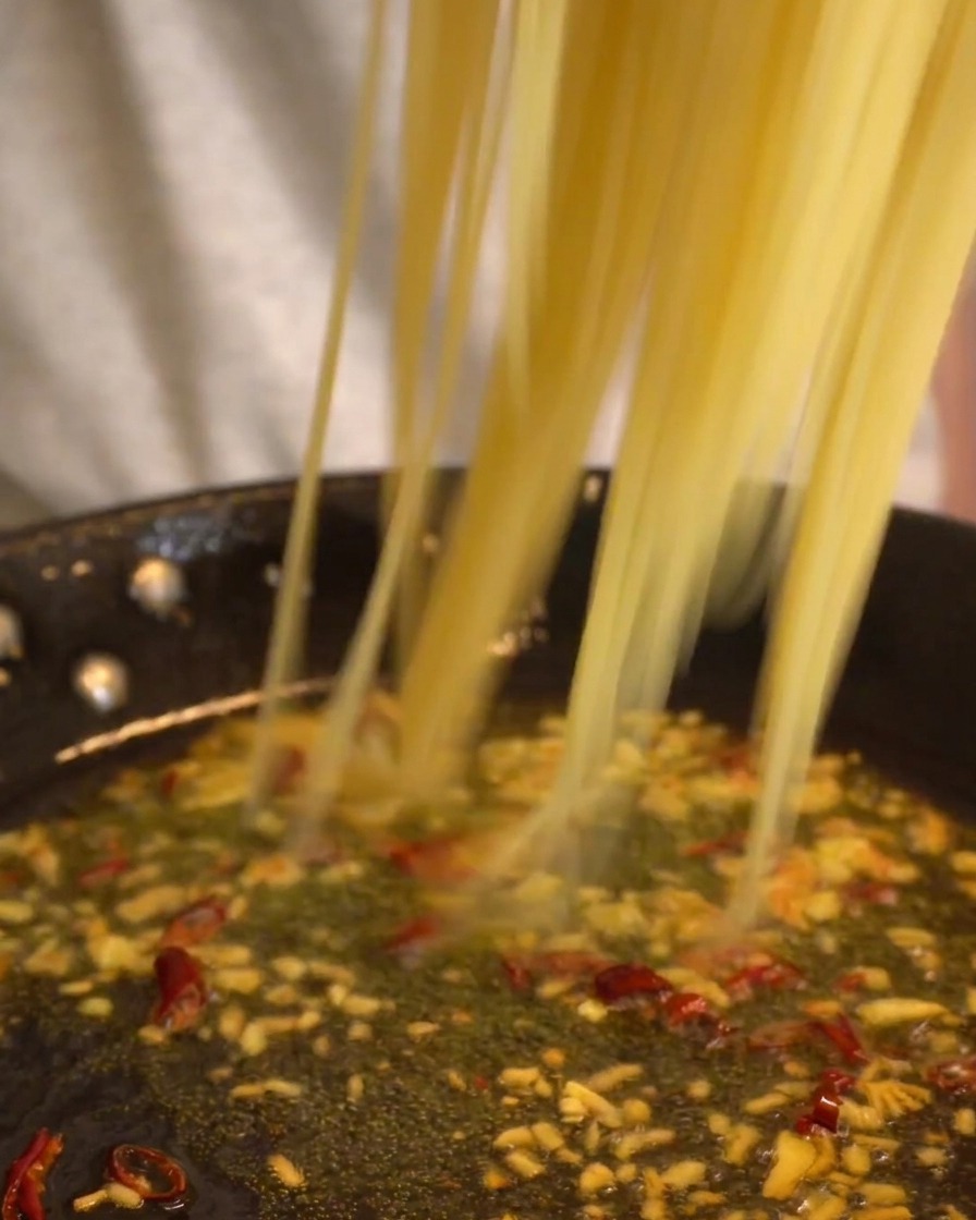 Espaguetis largos cayendo en una sartén con salsa de ajo y aceite.