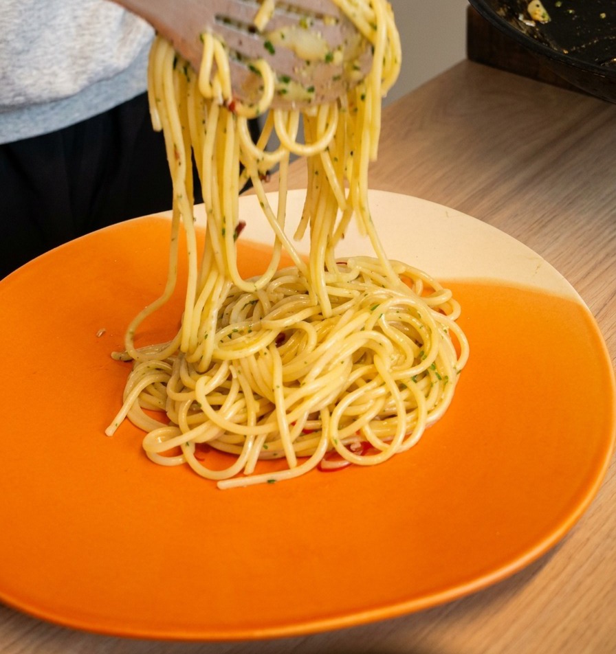Pinzas levantando y girando espaguetis aglio e olio sobre un plato naranja brillante.
