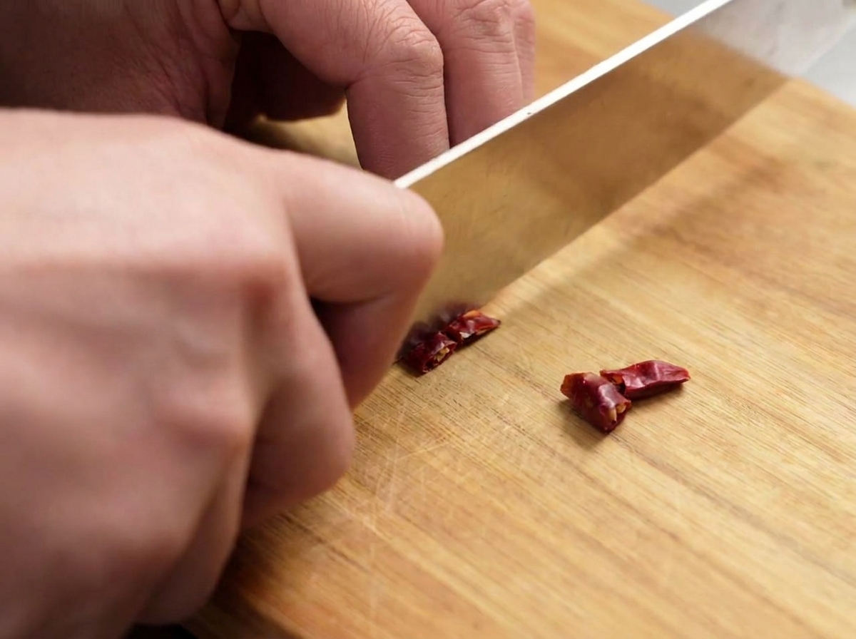 Un cuchillo cortando chiles rojos secos en trozos pequeños sobre una tabla.