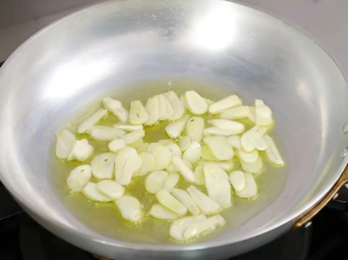 Láminas finas de ajo burbujeando suavemente en un fondo de aceite de oliva dentro de una sartén de metal.