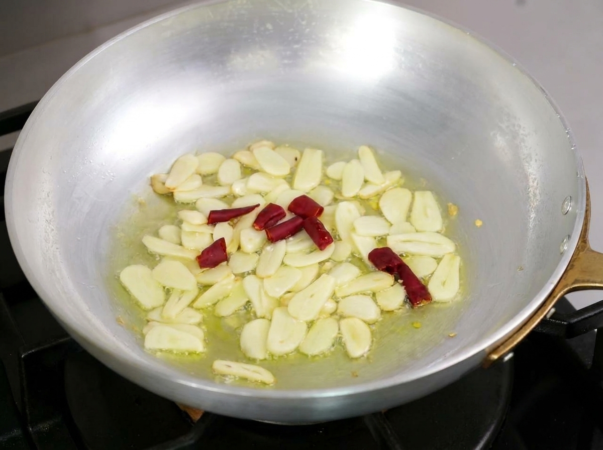 Ajo laminado y trozos de chile rojo seco burbujeando suavemente en aceite de oliva en una sartén.