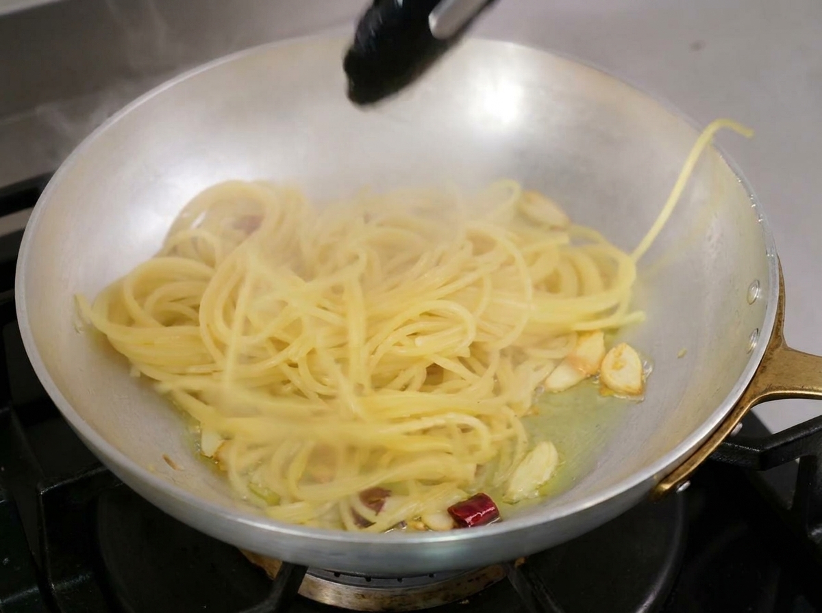 Espaguetis cocidos siendo trasladados con pinzas negras a una sartén con aceite infusionado con ajo y chile.