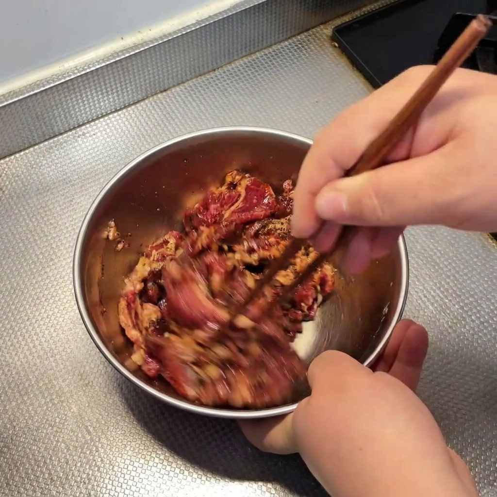 Manos usando palillos de madera para mezclar rápidamente la carne con el marinado en un bol metálico.