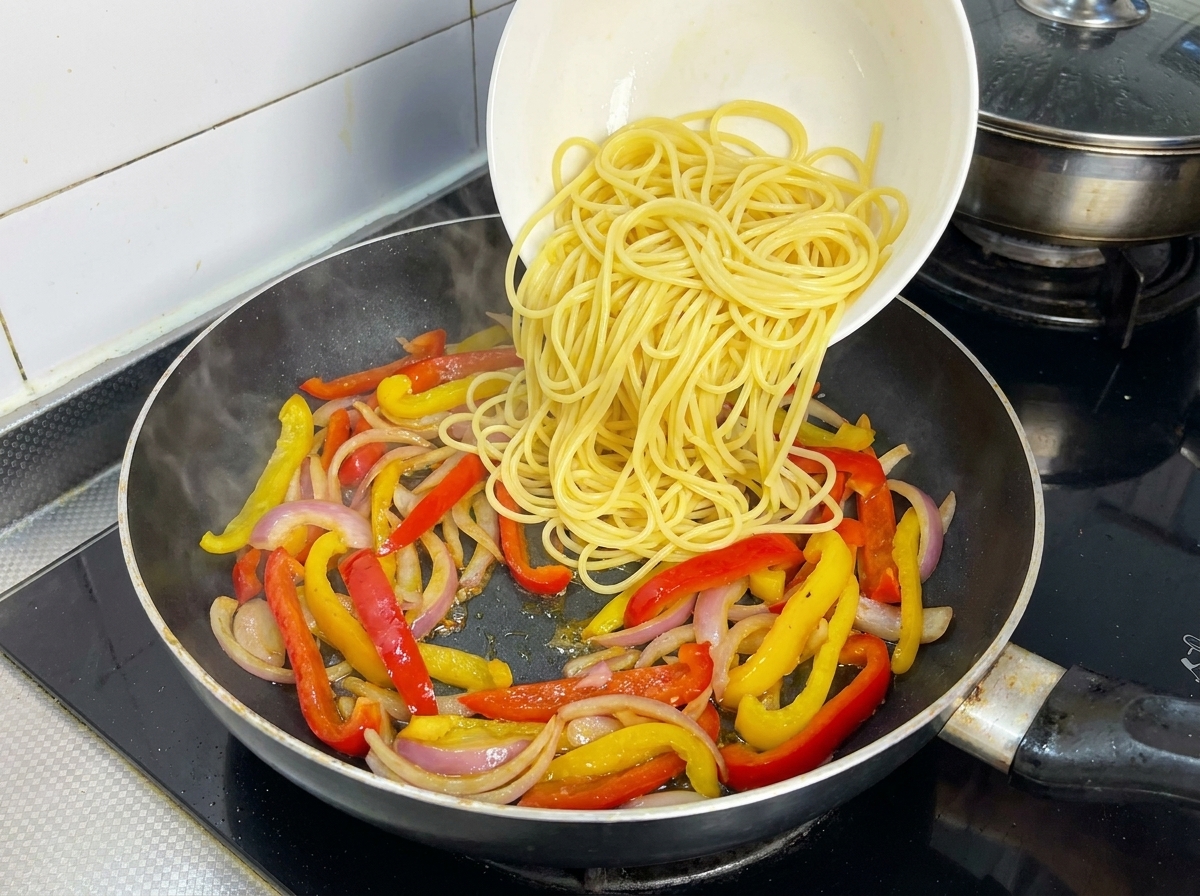 Espaguetis cocidos vertiéndose sobre la mezcla de pimientos y cebolla en la sartén.