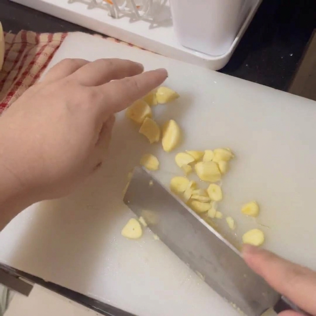 Manos usando un cuchillo para laminar finamente dientes de ajo frescos sobre una tabla blanca.