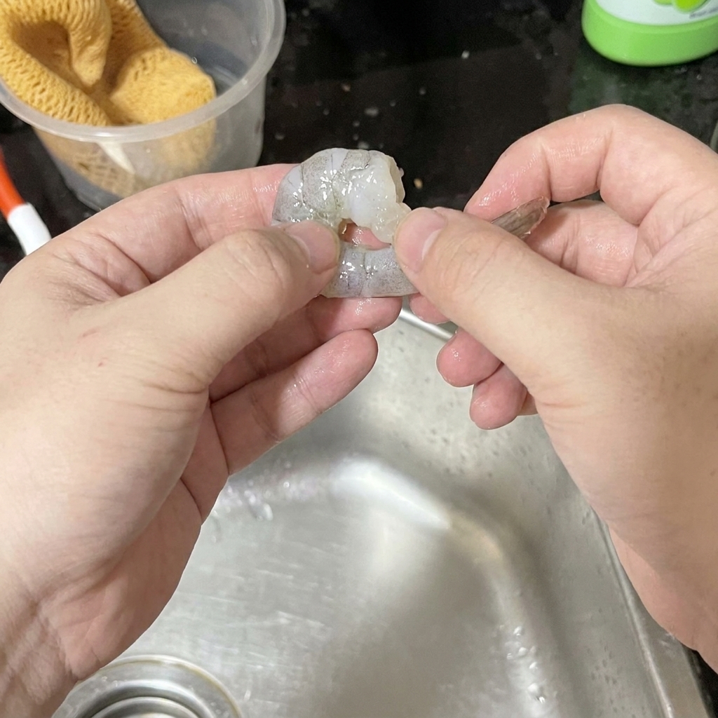 Dos manos pelando cuidadosamente un camarón crudo sobre un fregadero de acero inoxidable.