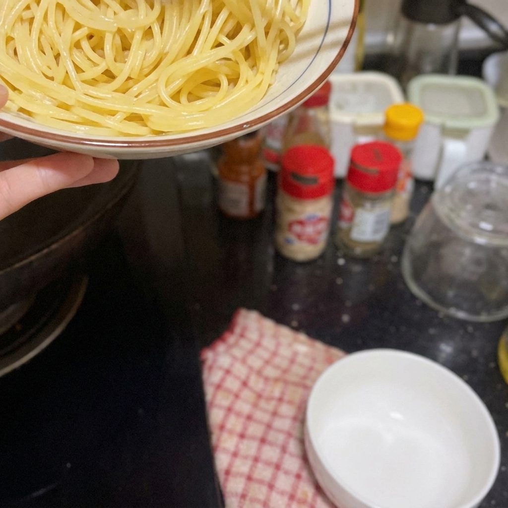 Una mano sosteniendo un bol de espaguetis cocidos sobre la estufa, listo para añadirlos a la sartén.
