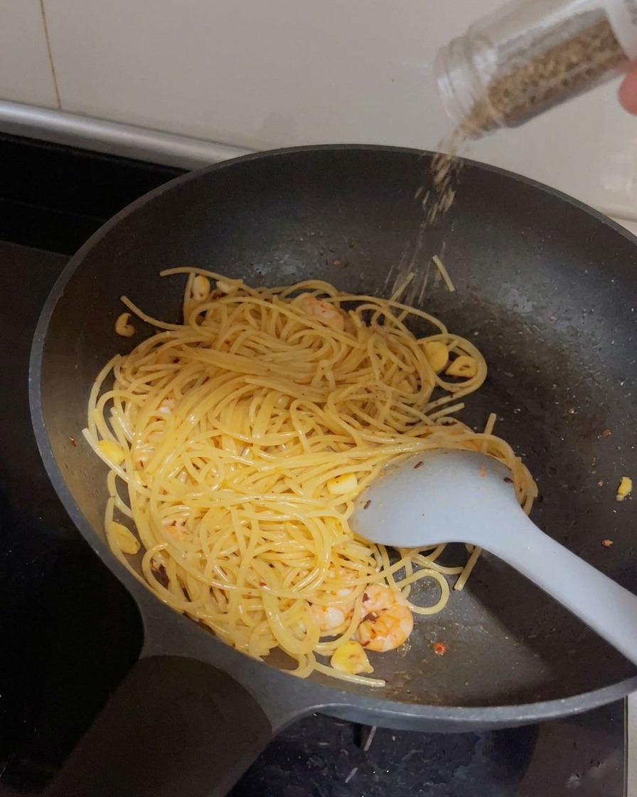 Un molinillo dispensando pimienta negra fresca sobre la sartén de espaguetis y camarones.