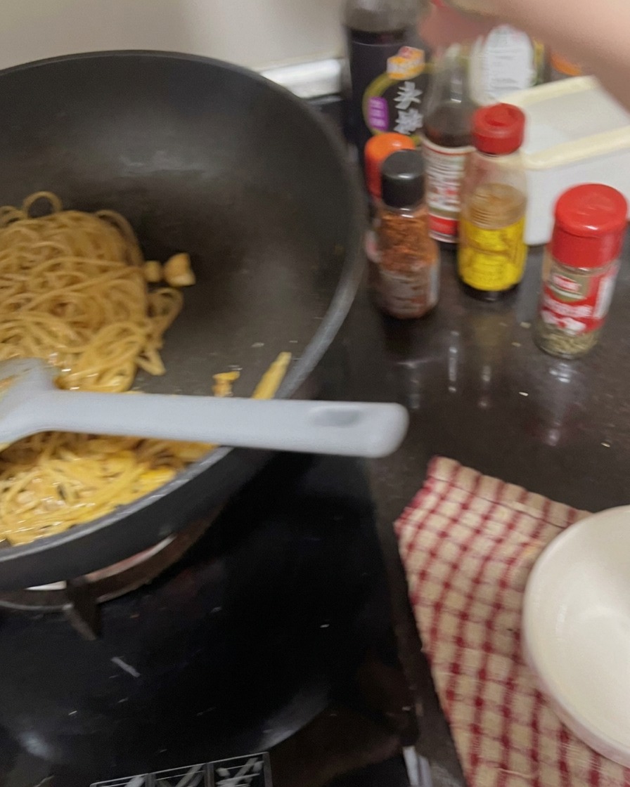 Una mano agitando un bote de especias sobre la sartén con fideos y ajo.
