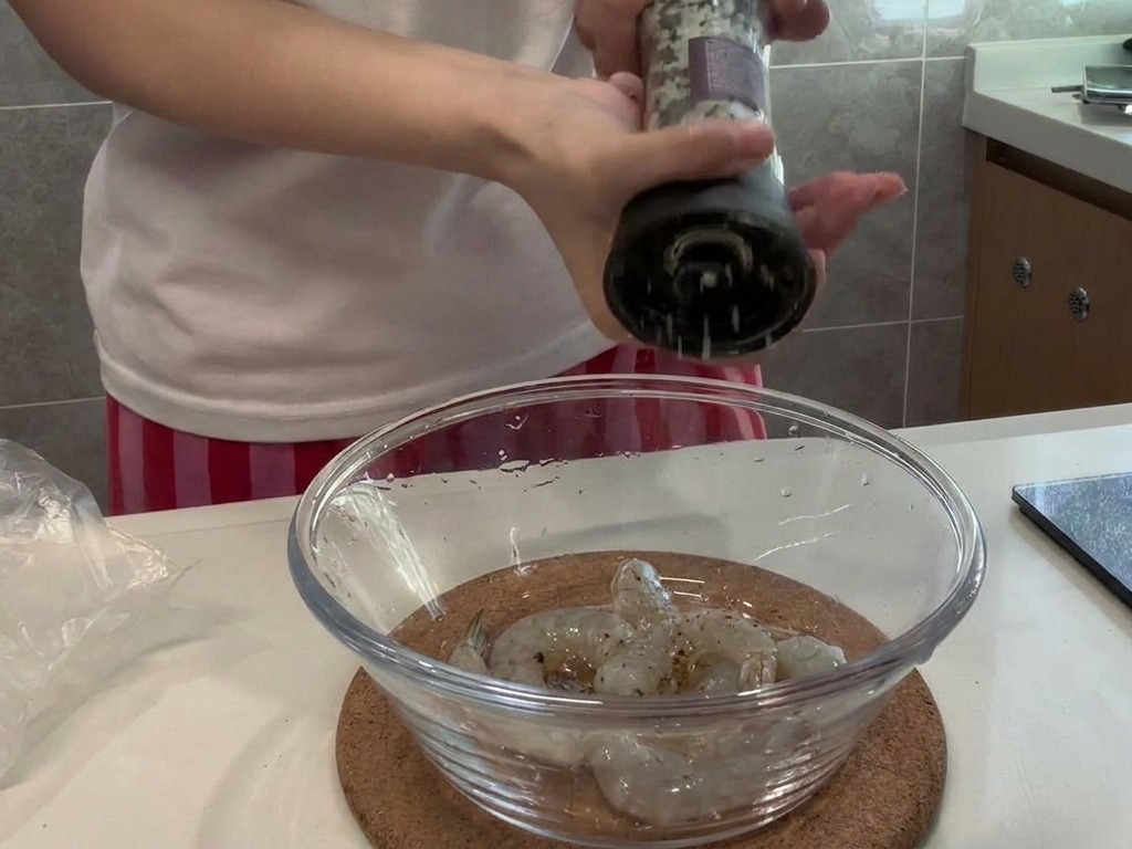 Pimienta negra recién molida cayendo sobre un cuenco de cristal con gambas crudas y peladas.