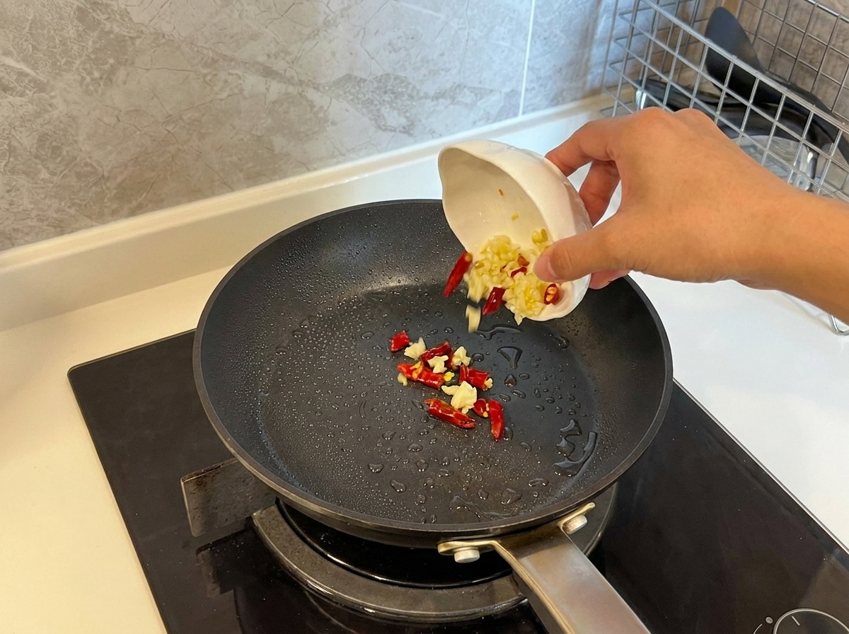 Ajo laminado y guindillas secas cayendo desde un pequeño cuenco a una sartén caliente con aceite.