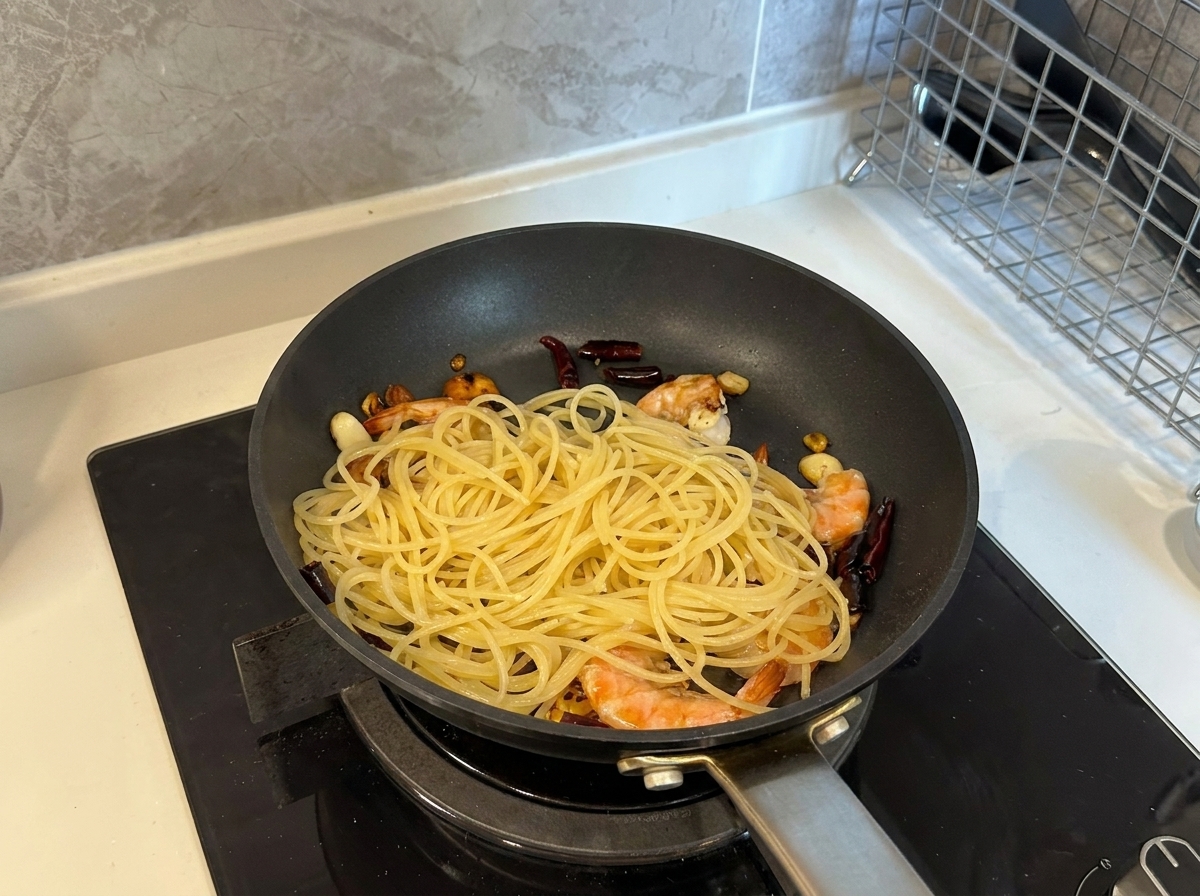 Espaguetis cocidos añadiéndose a una sartén llena de gambas cocinadas, ajo y guindillas.