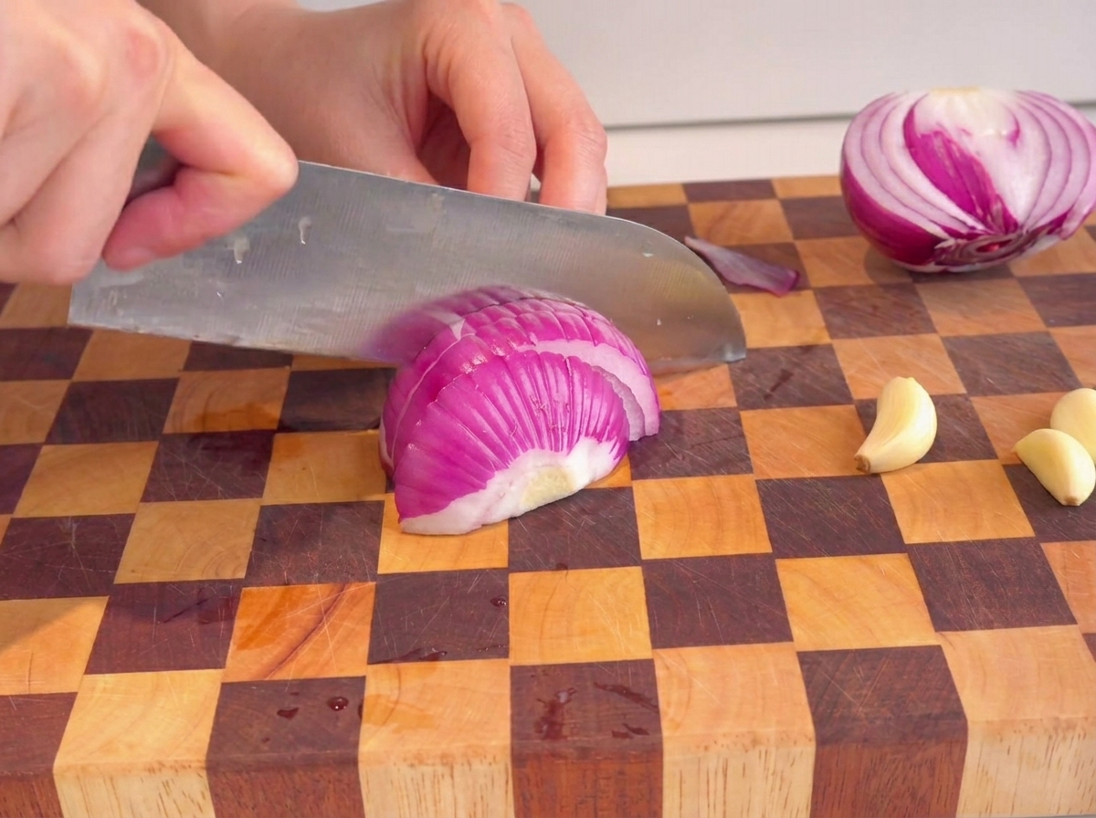 Manos usando un cuchillo para picar una cebolla morada sobre una tabla de cortar a cuadros.