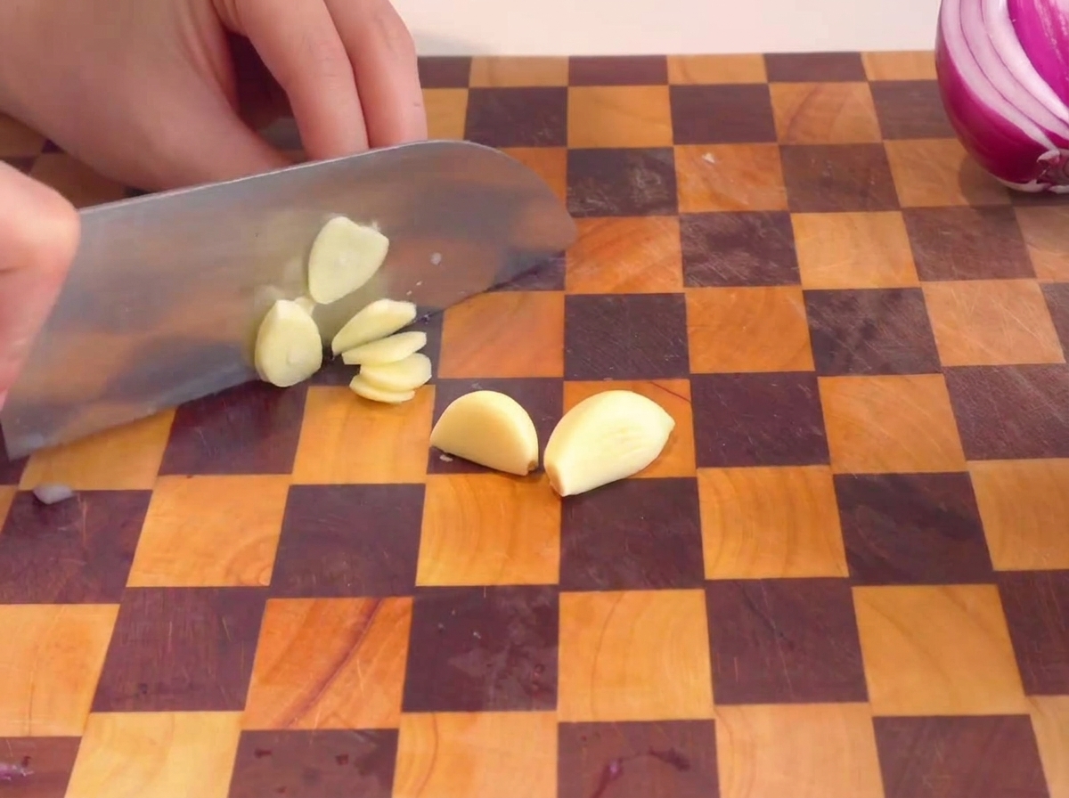 Manos laminando dientes de ajo pelados sobre una tabla de madera a cuadros.